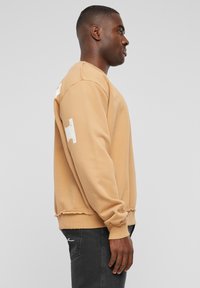 Khaki sweatshirt med rund halsudskæring, flagrende kant og hvide logo-accenter på ærmerne. Lavet af blødt bomuldsblandingsmateriale.