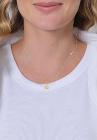 Collana d'oro con un pendente a forma di foglia di ginkgo texturizzata su una sottile catena attorcigliata, indossata sopra una maglietta bianca a coste.