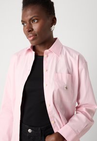 Ljust rosa knappad skjorta med en ficka och logodetalj, tillverkad av slät tyg. Bärs över en svart ribbad topp, matchad med mörka jeans.