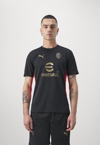 Black football shirt with red accents, featuring a textured pattern, gold logo, and Puma emblem. Worn with matching black shorts.