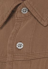 Camisa de mezclilla marrón con botones a presión plateados y detalle de costura. Presenta un cuello y dos cierres de botón en la parte delantera.