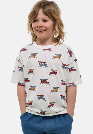 Enfant souriant aux cheveux blonds mi-longs, portant un t-shirt blanc orné de voitures de dessin animé colorées et un pantalon bleu.