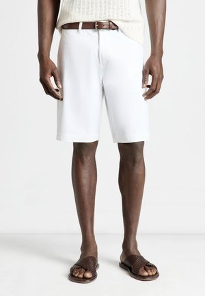 Uomo con pantaloncini bianchi al ginocchio, cintura marrone e sandali intrecciati marroni, in piedi su pavimento e sfondo bianchi.