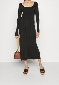 Donna che indossa un lungo vestito nero a costine con maniche lunghe e scollo quadrato, tiene in mano una borsa marrone e indossa sandali neri con plateau.