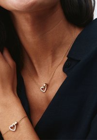 Moteris dėvi auksinį širdies formos kaklo papuošalą ir apyrankę su mažais deimantais, apsirengusi tamsiai mėlyna marškinėlių su apykakle.