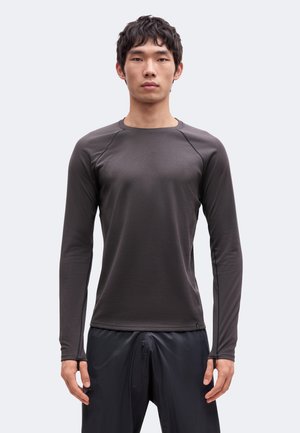 Mann, der nach vorne schaut, trägt ein eng anliegendes dunkelgraues Langarmshirt und lockere schwarze Hose vor einem schlichten hellen Hintergrund.