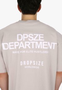 Világosszürke, túlnőtt póló, melynek hátulján feltűnő fehér felirat található: "DPSZE DEPARTMENT," "MADE FOR ELITE HUSTLERS," és "DROPSIZE WORLDWIDE."