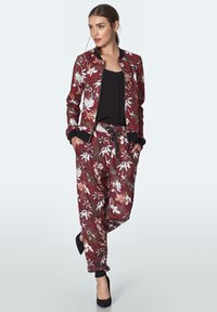 Kobieta w bordowej, kwiecistej bomberce i pasujących spodniach, z czarną bluzką i czarnymi szpilkami, stojąca z rękami w kieszeniach na jednolitym tle.