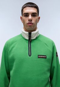 Groene fleece trui met een hoge kraag, een halve ritssluiting aan de voorkant en een contrasterende witte binnenkant. Voorzien van een geborduurd logo op de borst.