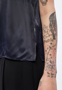 Tmavomodrý saténový top s rovnomerným leмом, bočnými rozparkami a malou kovovou ozdobou. Koža s detailnými tetovaniami na ruke je viditeľná.