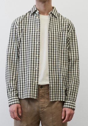 Hombre con camisa de cuadros en blanco y negro desabrochada sobre una camiseta blanca y pantalones marrones, con un fondo claro liso.