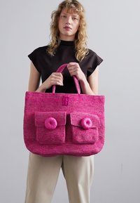 Bolsa de tote tejida en fucsia con dos bolsillos frontales y acentos de botón. Cuenta con asas redondeadas y resistentes, y un acabado texturizado.