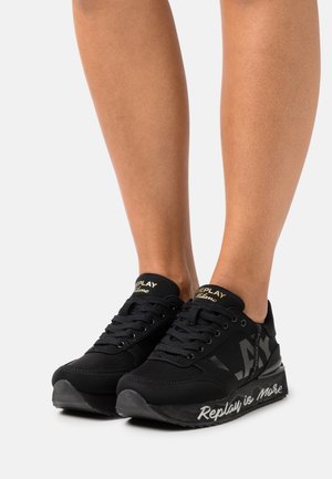 Baskets noires avec une tige en mesh et synthétique, dotées d'une semelle surélevée. Les accents comprennent un logo doré et un détail répétitif "Replay is More".
