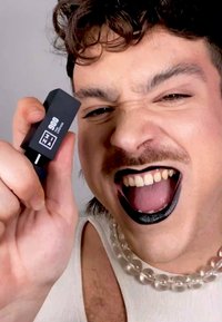 Svart leppestiftube med hvit etikett holdt i en hånd. Subjektet har svart leppestift på leppene, iført en perlekjede og hvit tanktop.