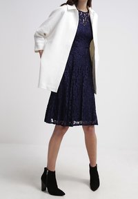 Navy lace dress with fitted bodice and flared skirt, layered under a white oversized coat. Black ankle boots complete the look.