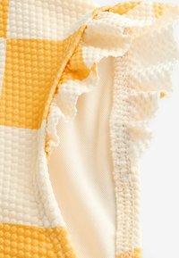 Tissu à carreaux jaunes et blancs avec une surface texturée ; présente des bords froncés et une doublure lisse ; détail en gros plan de la construction du vêtement.