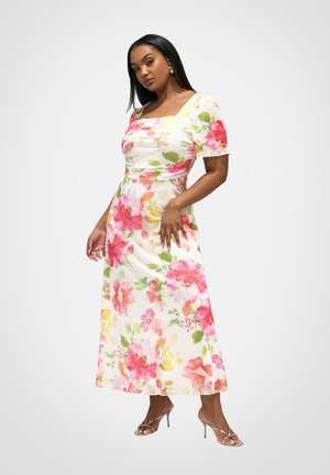 Frau trägt ein weißes, geblümtes Midikleid mit pinken und grünen Blumen, quadratischem Ausschnitt, kurzen Ärmeln und beigen Sandalen mit Absatz, posiert vor weißem Hintergrund.