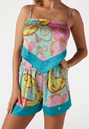 Femme portant un haut bustier coloré et un short assorti avec un imprimé chaîne et ceinture, bordures turquoise, et une main sur la hanche.