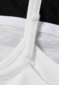 Sangle de soutien-gorge blanche avec matériel ajustable, reposant sur un tissu superposé gris et noir. Le soutien-gorge présente une texture lisse et une couture détaillée.