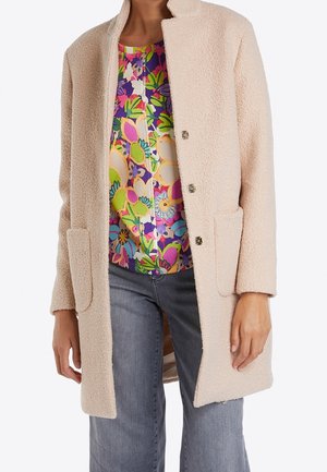 Manteau en laine beige texturé avec des poches avant, porté ouvert sur un chemisier fleuri multicolore vif et un jean gris.