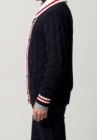 Marinblå kabelstickad cardigan med vit krage och röda accenter. Har knäppning med knappar, två framfickor och randiga ärmslut.