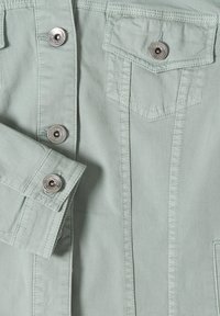 Světlý zelený úsek bundy s kovovými knoflíky, přední kapsou s klopou a detailním prošitím na manžetě a švech.