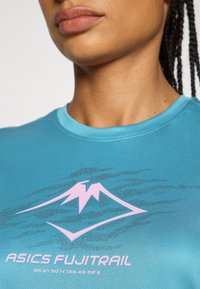 Turquoise athletic shirt featuring a pink mountain logo and text "ASICS FUJITRAIL," with a smooth, textured fabric. Round neckline.