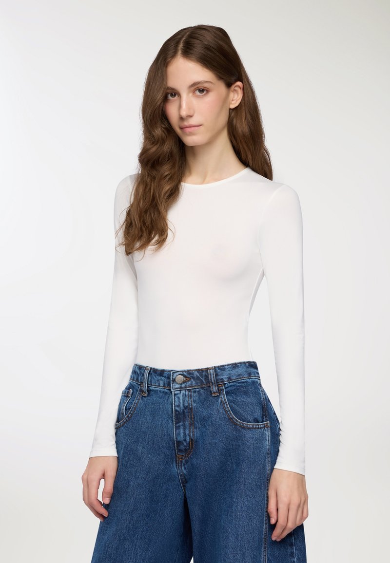 Maglietta bianca a maniche lunghe abbinata a pantaloni larghi in denim blu scuro a vita alta. Tessuto liscio, scollatura rotonda e dettagli di cucitura visibili.