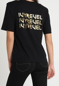 Černé tričko s kovově zlatým textem "IVYREVEL" na zadní straně, krátké rukávy, kulatý výstřih a uvolněný střih.