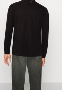 Schwarzes Langarmshirt mit rundem Halsausschnitt, glatter Textur und dezenten Details im Rücken, kombiniert mit lockeren, dunkelgrauen Jeans.