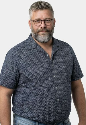 Mittelalter Mann mit grauem Bart und Brille, der ein dunkles gemustertes kurzärmeliges Hemd und hellblaue Jeans trägt, steht vor einem schlichten Hintergrund.