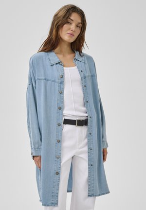 Mladá žena v dlhom svetlom modrom rifľovom kabáte na gombíky, oblečená cez biely tielko a biele nohavice s čiernym opaskom, na jednoduchom pozadí.