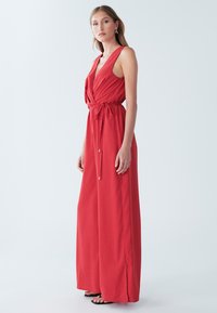 Rode jumpsuit met een wikkeldesign, zonder mouwen, met een trekkoord in de taille en vloeiende stof, getoond op een model tegen een lichte achtergrond.