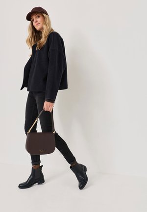 Pull à col montant en polaire noire, jeans skinny noirs, bottes Chelsea noires et un sac à main structuré marron avec une bandoulière dorée. La mannequin pose de manière dynamique.