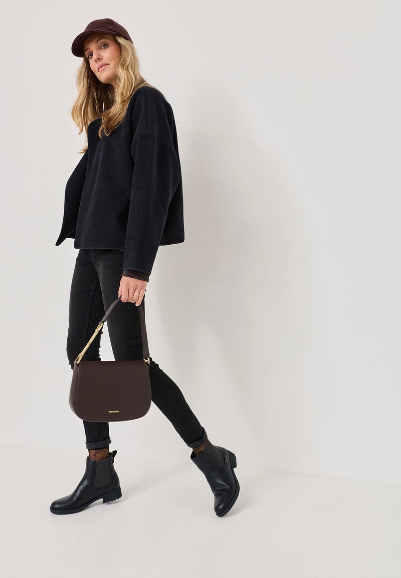 Pulover negru din fleece, blugi negri skinny, cizme negre până la glezne și o geantă structurată maro cu o curea aurie. Modelul pozează dinamic.