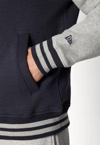 Sudadera de tiro navy con mangas grises, que cuenta con un bolsillo frontal, puños acanalados con rayas grises y blancas, y un detalle de logo en la manga.