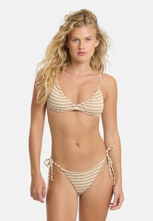 Blondynka o falowanych włosach, ubrana w beżowo-biały paskowany bikini z cienkimi ramiączkami i wiązaniami po bokach, stojąca na jednolitym jasnym tle.