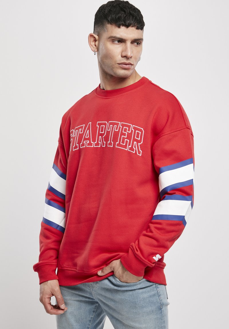 Roter Sweatshirt mit "STARTER" in Weiß gedruckt. Besitzt blaue und weiße Streifen an den Ärmeln, gerippte Bündchen und einen Rundhalsausschnitt.