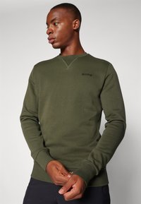 Sudadera verde oliva hecha de tela suave, con cuello redondo, puños de canalé y un sutil logo negro en el pecho.