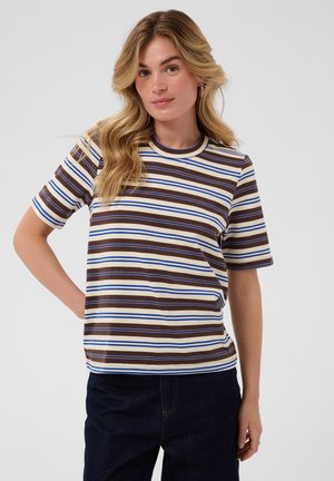 Femme aux cheveux blonds ondulés porte un t-shirt à manches courtes rayé marron, blanc et bleu et un pantalon foncé, posant une main sur la hanche.