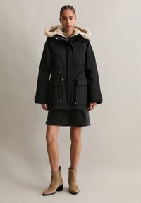 Zwarte parka met een beige faux fur gevoerde capuchon, voorkruiszakken en een verstelbare taille. Gecombineerd met een donkerdenim rok en beige enkellaarsjes.