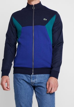 Mężczyzna w kurtce na suwak w kolorach niebieskim, morskim i granatowym z małym logo krokodyla na piersi oraz niebieskich dżinsach.