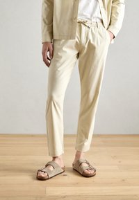 Beżowe spodnie z bawełny z domieszką, z elastycznym pasem i dopasowanym krojem, w połączeniu z szarymi zamszowymi sandałami z podwójnymi paskami i korkową podeszwą.