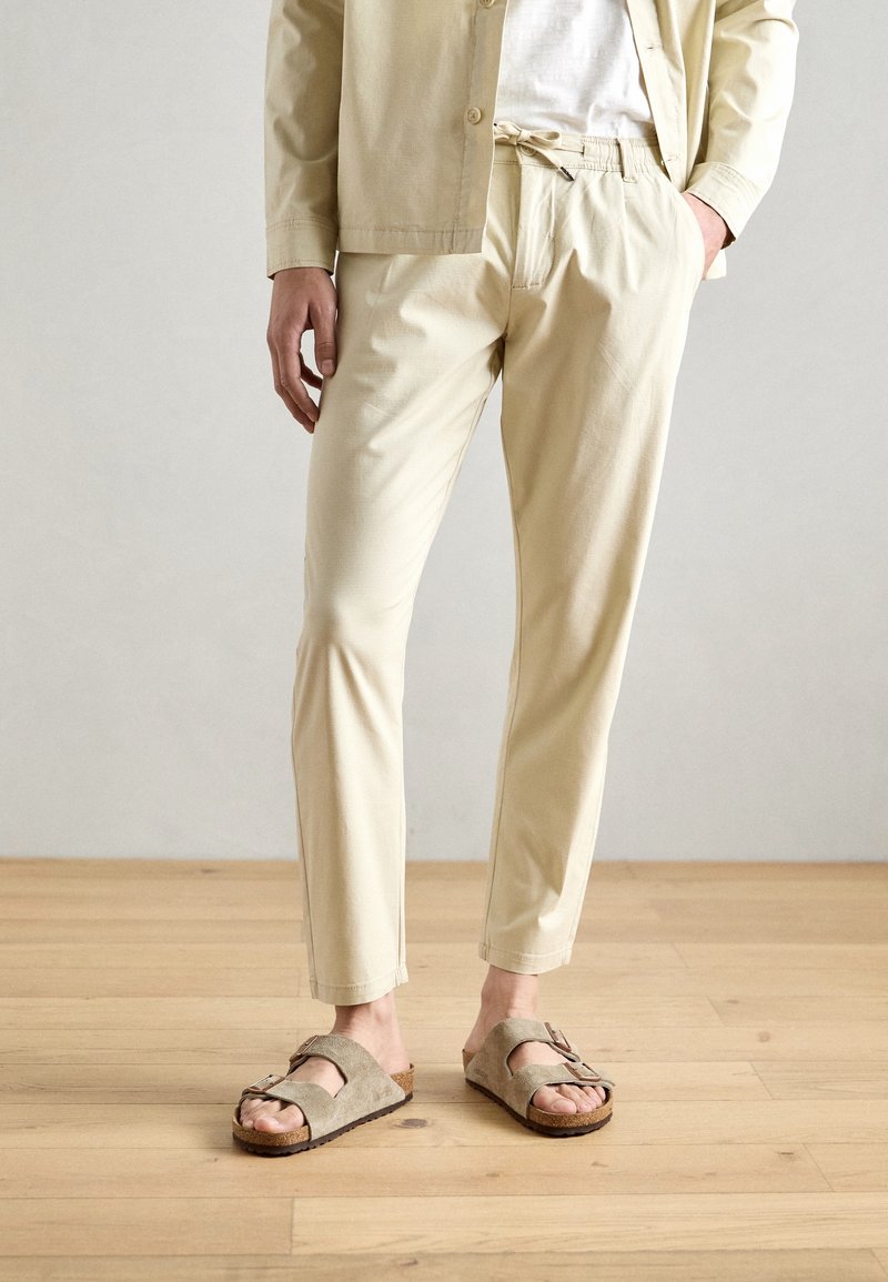Beżowe spodnie z bawełny z domieszką, z elastycznym pasem i dopasowanym krojem, w połączeniu z szarymi zamszowymi sandałami z podwójnymi paskami i korkową podeszwą.