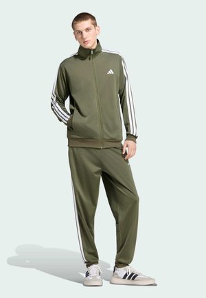 Junger Mann trägt olivgrünen Adidas Trainingsanzug mit weißen Streifen und weißen Adidas Sneakern, steht mit einer Hand in der Tasche.