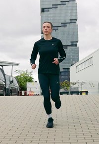 Woman in black athletic wear running on a paved path with modern glass building and parked vehicles in the background.