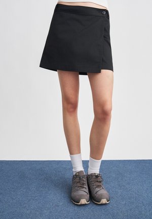 Fekete wrap szoknyát, fehér bokazoknit és szürke túracipőt viselő személy kék szőnyegen áll, fehér fal előtt.