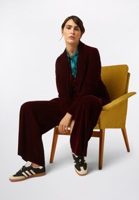 Traje de terciopelo burdeos con pantalones de pierna ancha, camisa teal con estampado y zapatos negros con detalles en crema, todo sobre una silla amarilla. Fondo sencillo.
