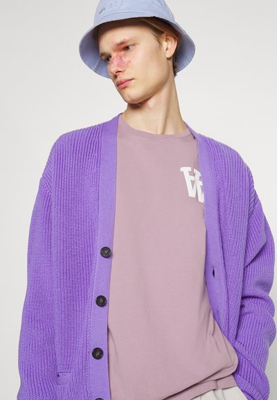 Violetiniu megztu kardiganu su raukšlėta tekstūra, su mygtukų uždarymu, dėvimas virš šviesiai violetinio marškinėlių su balta logo. Šviesiai mėlyna kibiro skrybėlė.