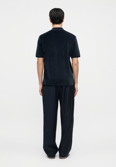 Κοντομάνικο πόλο μπλουζάκι σε ναυτικό μπλε με απαλή υφή, συνδυασμένο με χαλαρό παντελόνι με λεπτές ρίγες, εμφανιζόμενο από πίσω πάνω σε λευκό φόντο.